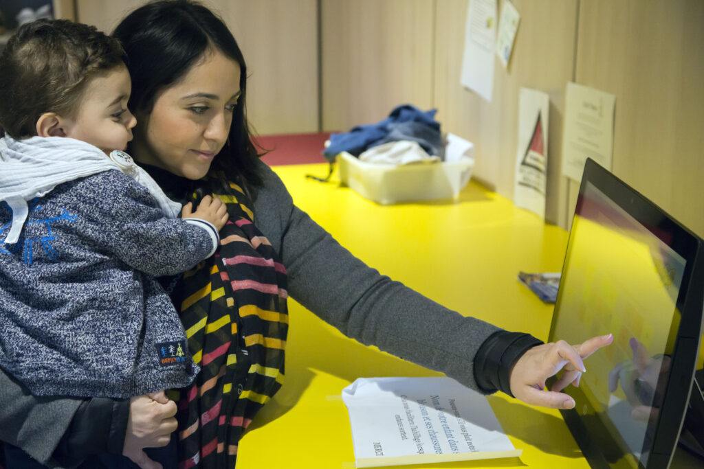 Une jeune femme tient son enfant dans les bras et clique sur un écran tactile